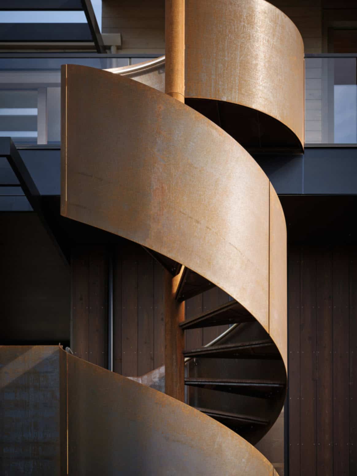Corten steel spiral staircase framed by sunlight and dark siding