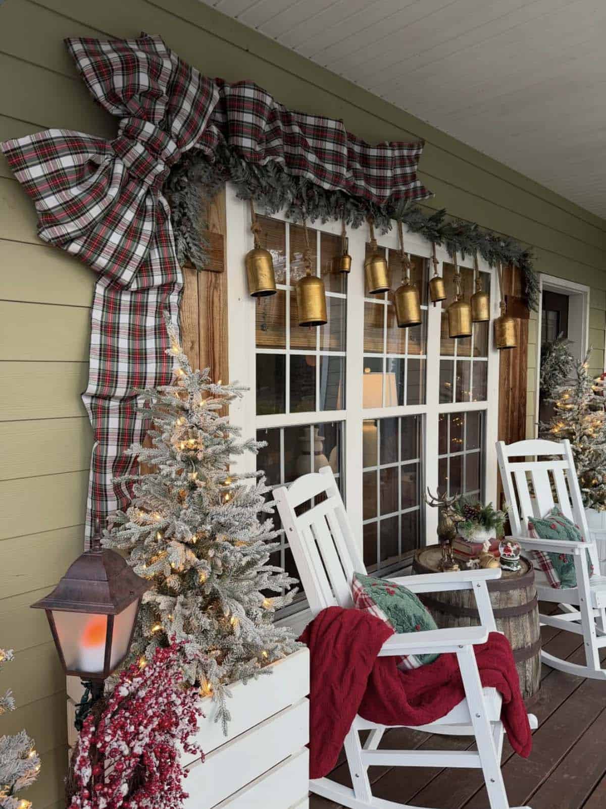 Christmas porch with an oversized tartan plaid bow and evergreen garland framing a window, brass bells, flocked trees, and white rocking chairs