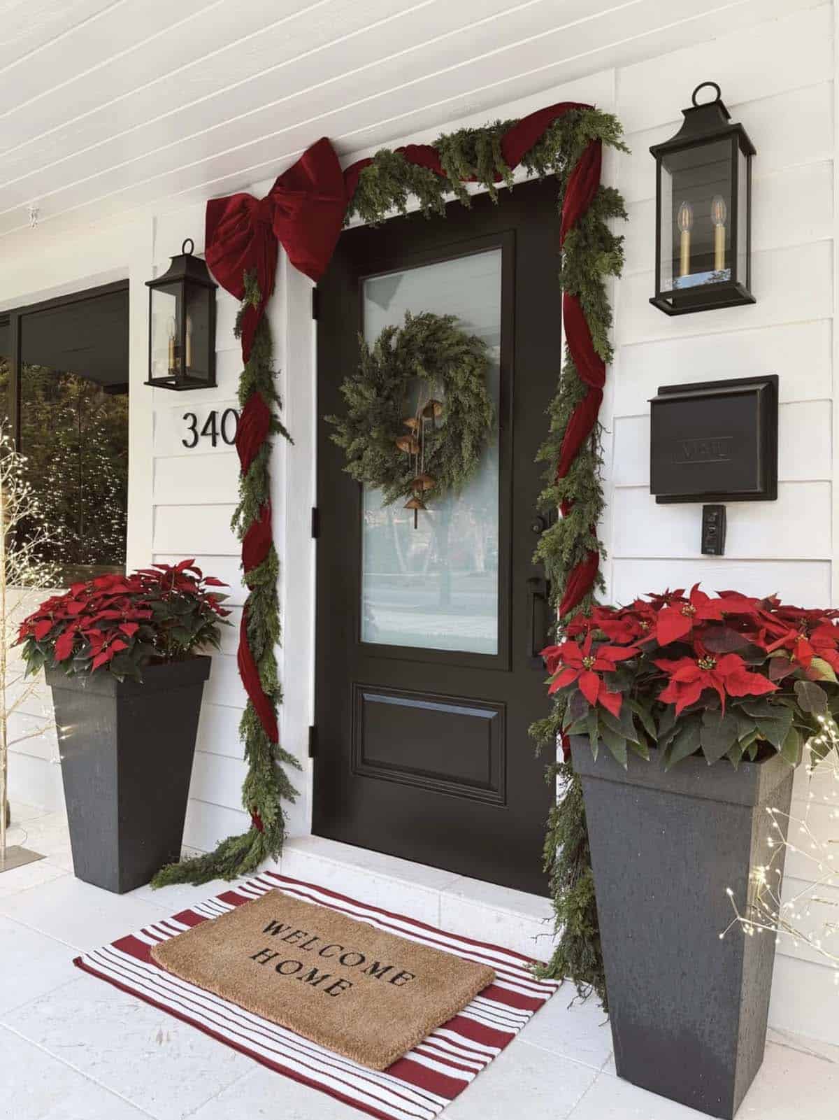 Black front door framed by an evergreen garland wrapped in deep red velvet ribbon with an oversized bow, flanked by tall planters of red poinsettias