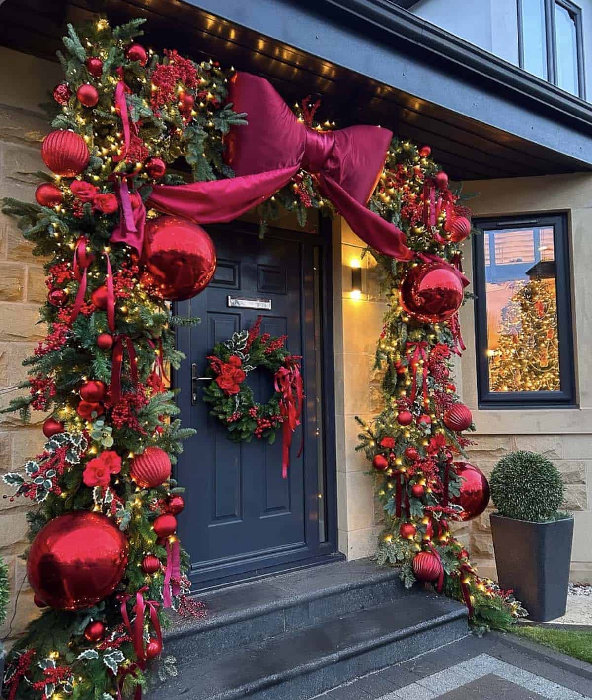 Front door framed by an evergreen garland with a giant red satin bow, oversized red ornaments, berries, and twinkle lights
