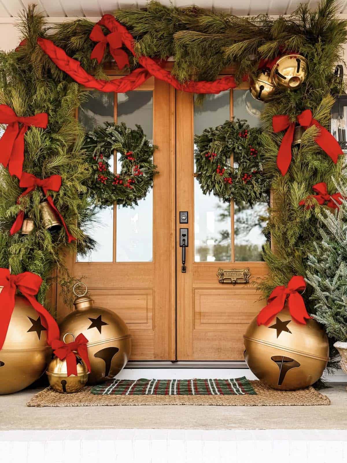 Wood double front doors framed by a pine garland with red bows, flanked by giant gold jingle bells, with evergreen wreaths on each door