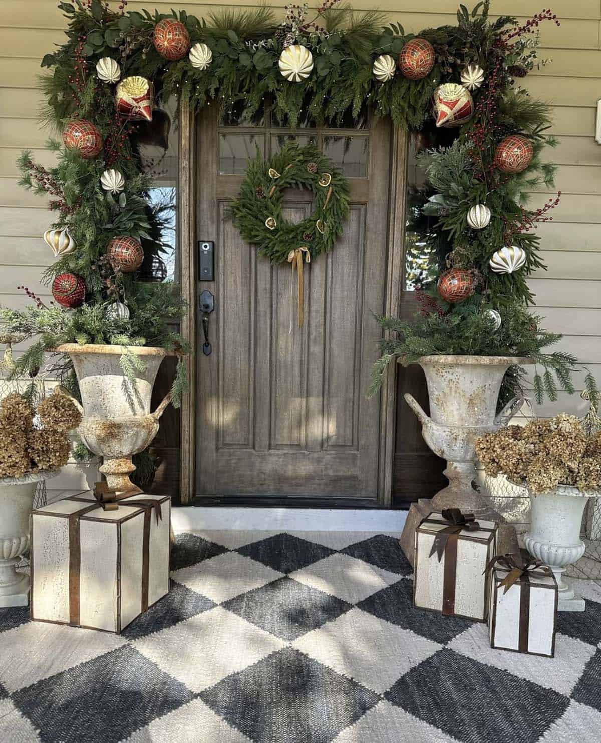 Wood front door framed by an evergreen garland with red plaid and white ornaments, flanked by weathered iron urns, dried hydrangeas, and metal gift boxes on a black and white harlequin rug