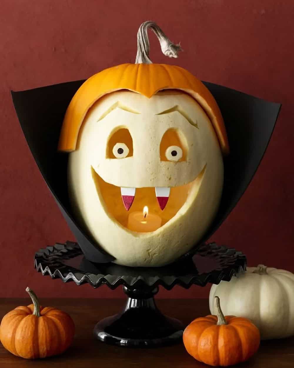 White pumpkin carved as a smiling vampire with wooden fangs, an orange widow’s peak, and a black foam collar