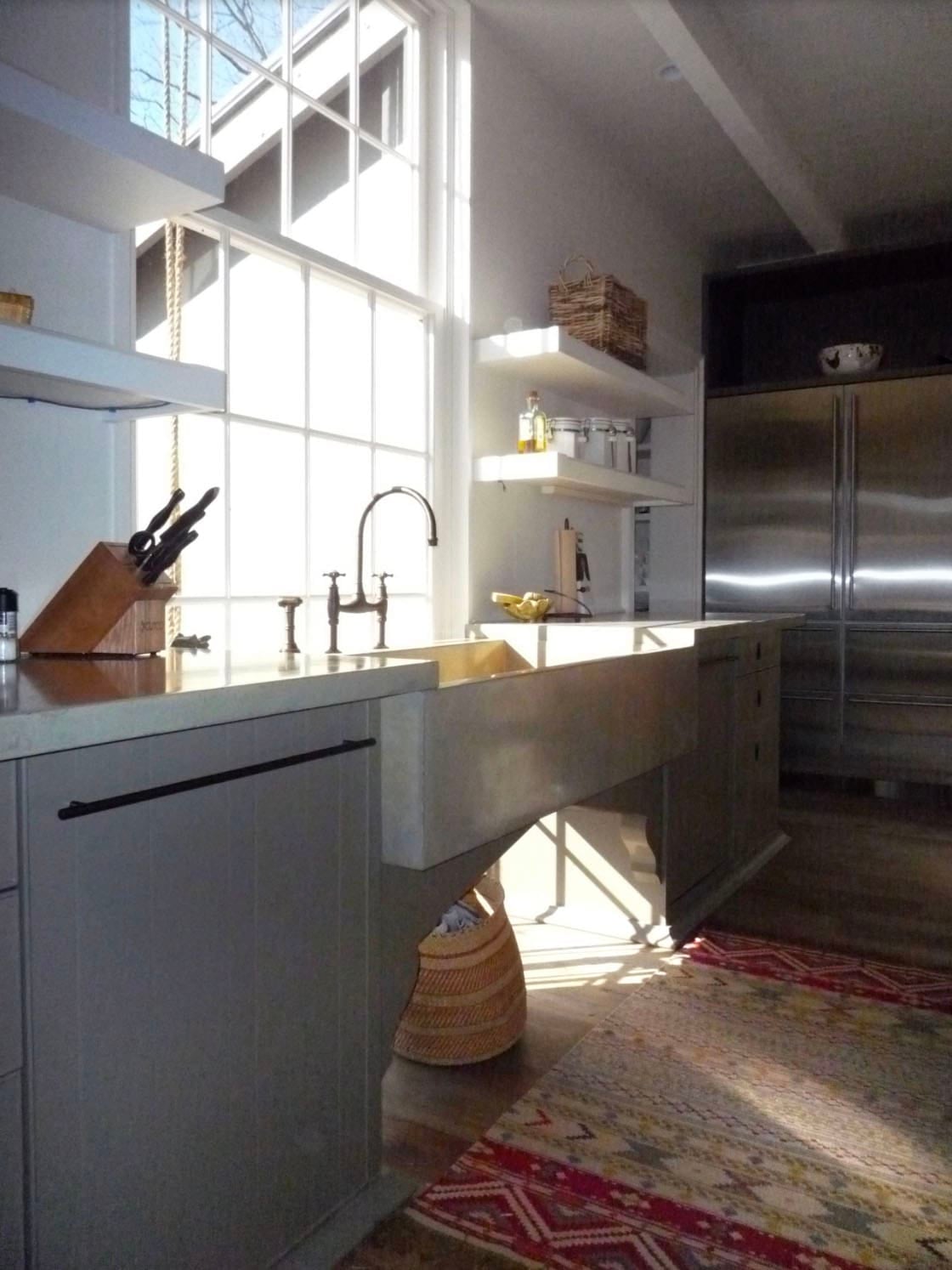 Concrete farmhouse sink beneath a tall window, with gray cabinets and open white shelves