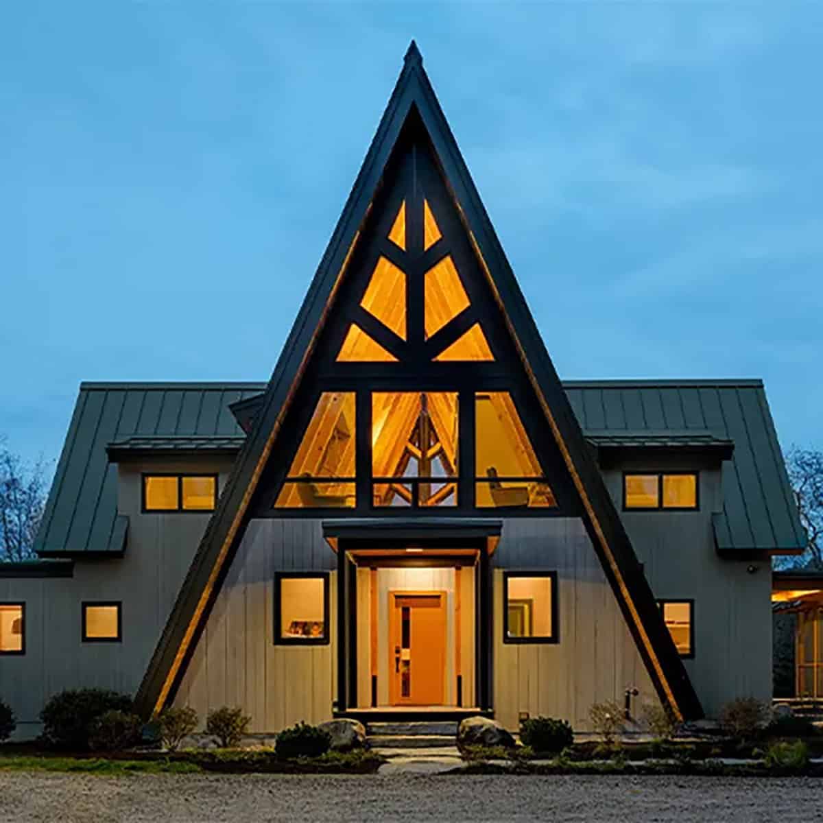 A-frame mountain home at dusk with a glowing triangular window wall, dark standing seam metal roof and light vertical wood siding