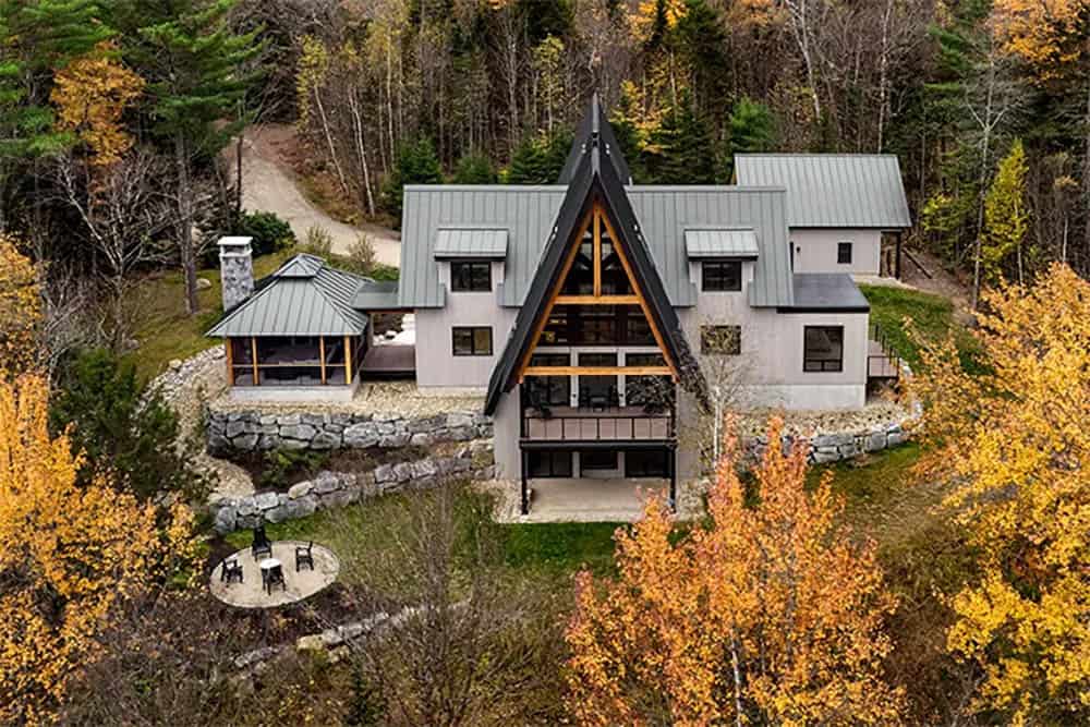 Aerial view of the A-frame home in fall woods, with stone retaining walls, a screened porch and a round fire pit patio