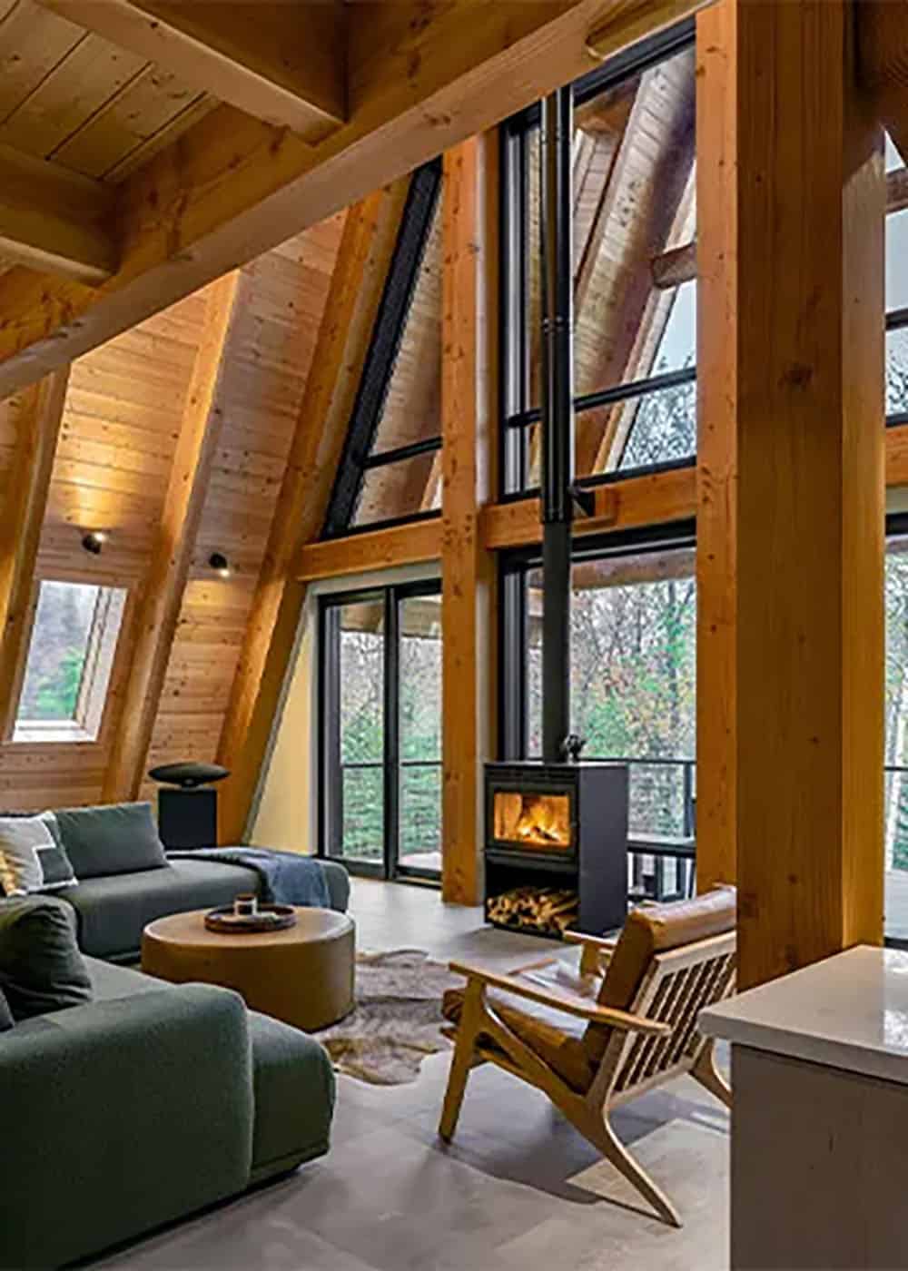 Living room with a green sectional, a black wood stove and two-story windows set into exposed timber framing