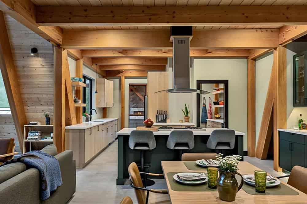 Open kitchen with light wood cabinets, a dark green island with gray stools and a stainless hood under timber beams