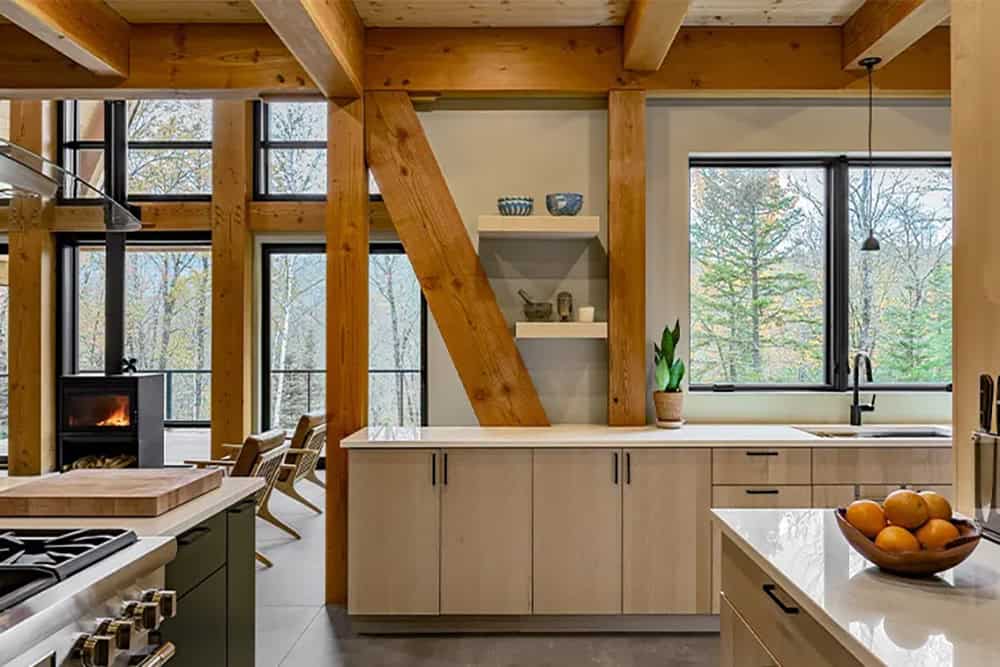Kitchen counter with light maple cabinets between a diagonal timber brace and post, with the wood stove and windows beyond