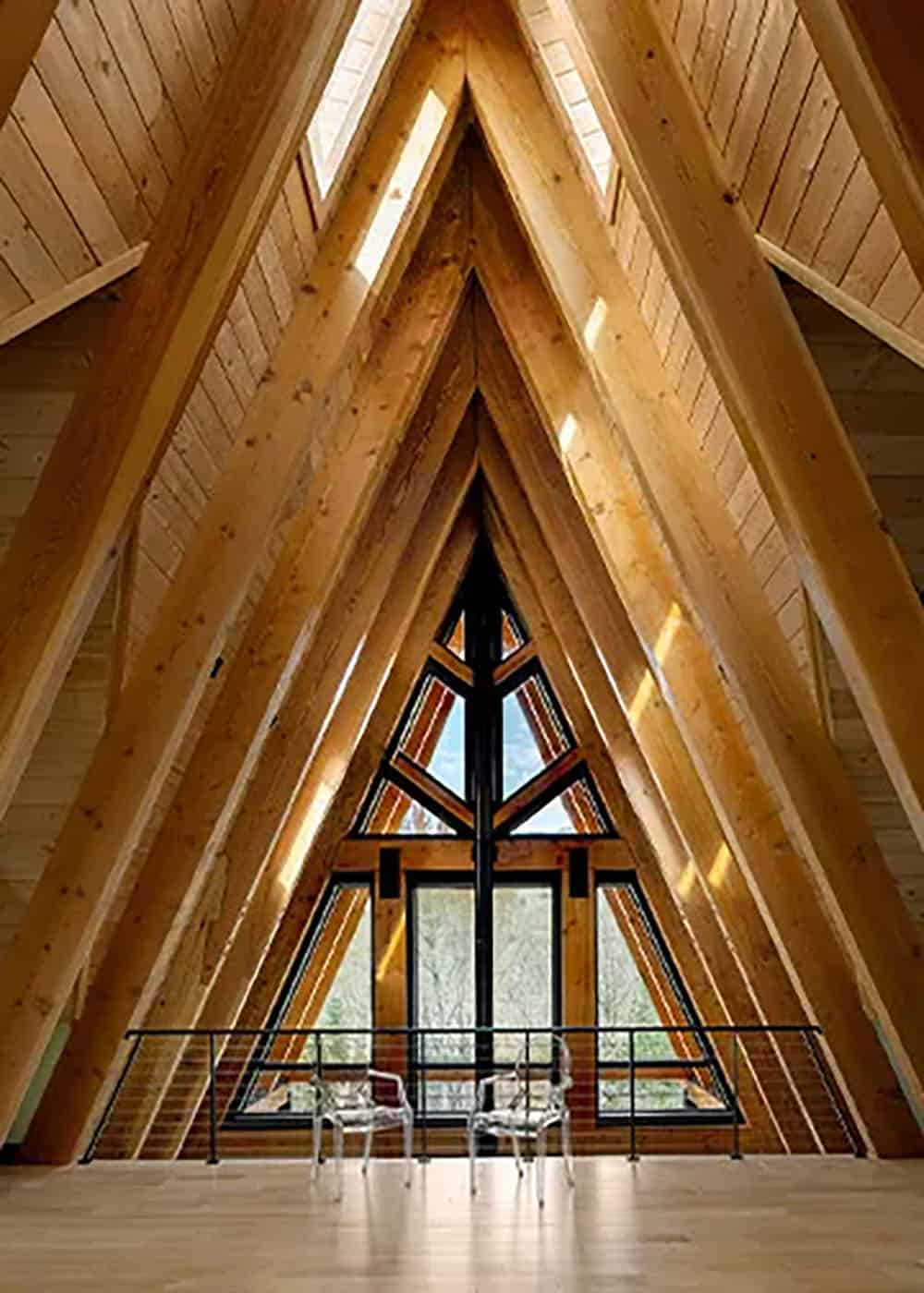 Loft under rows of timber rafters facing a tall triangular window wall, with two clear acrylic chairs at the railing