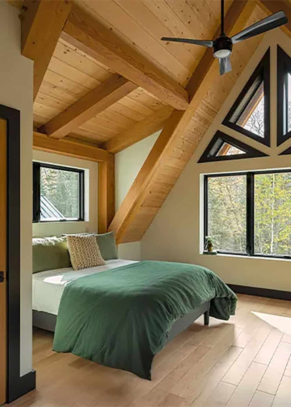 Bedroom with green bedding under a sloped timber ceiling, with triangular windows above a wide window to the woods