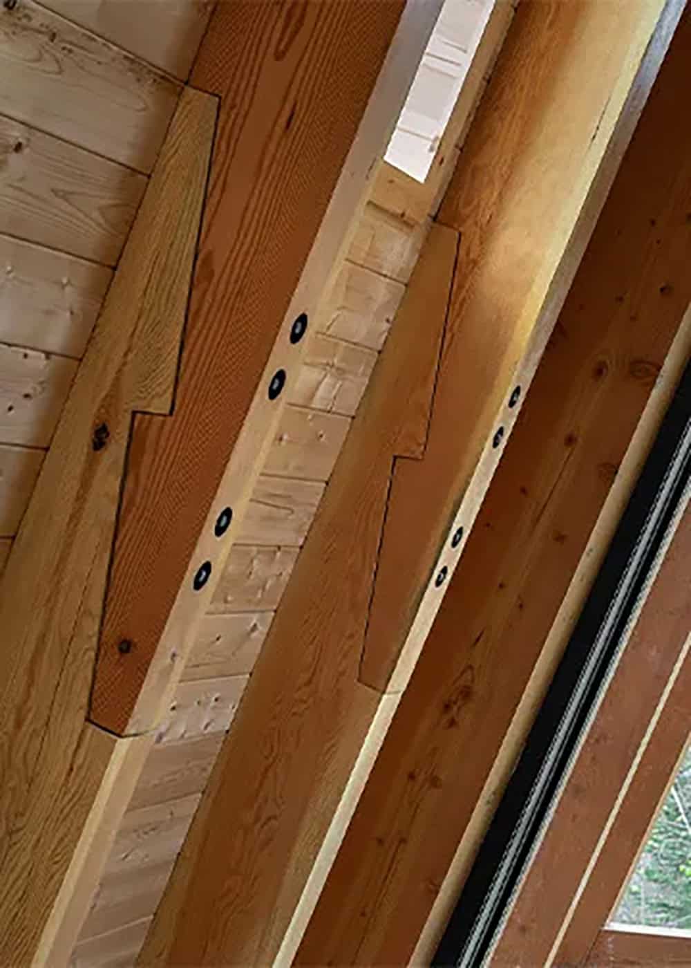 Close-up of timber frame joinery with notched rafters and black bolt plates against a pine plank ceiling