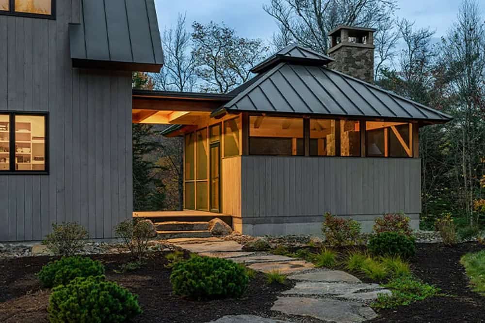 Screened porch at dusk with a hipped metal roof and stone chimney, reached by a flagstone path through landscaping