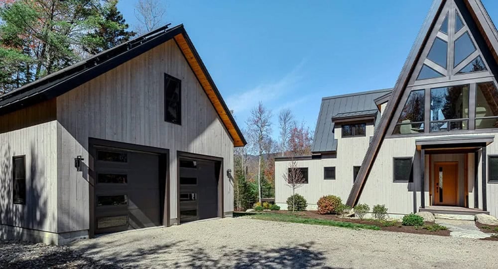 Detached garage with black doors across a gravel drive from the A-frame home's glass gable and front door
