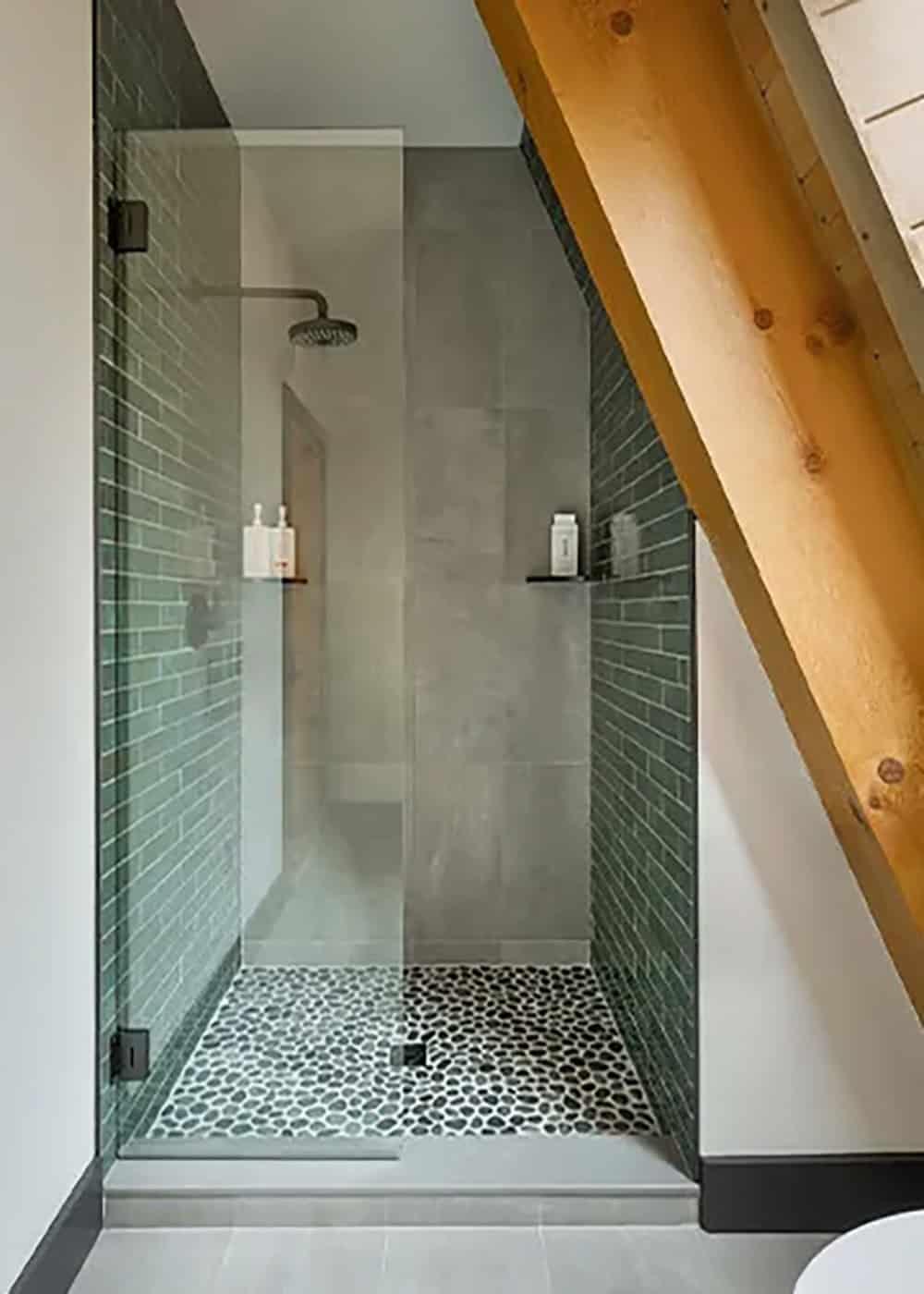 Glass walk-in shower with green brick-style tile, gray tile walls and a pebble floor under a sloped timber rafter