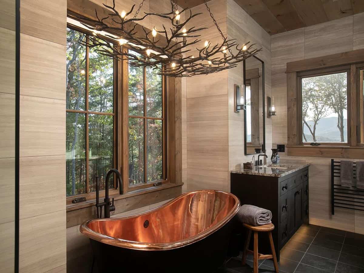 Copper soaking tub under a branch chandelier beside wood framed windows
