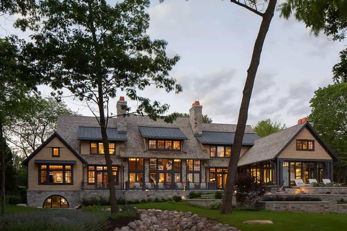 Lakeside elevation with metal-roofed dormers, stone chimneys and lit windows at dusk