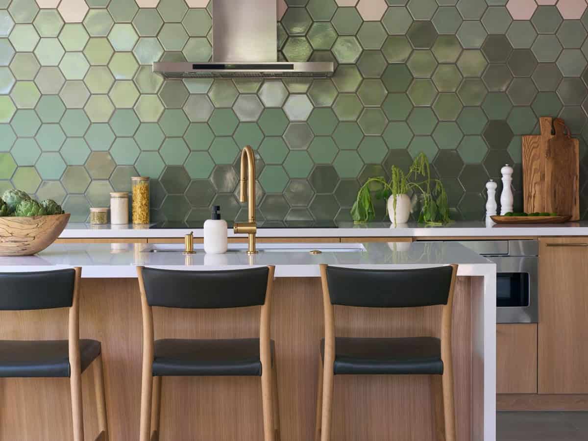 Brass faucet on a white quartz island in front of a glossy green hexagon tile backsplash and stainless range hood