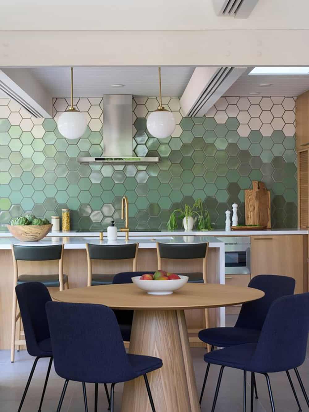 Round oak pedestal dining table with navy upholstered chairs in front of the kitchen island and green hexagon tile wall
