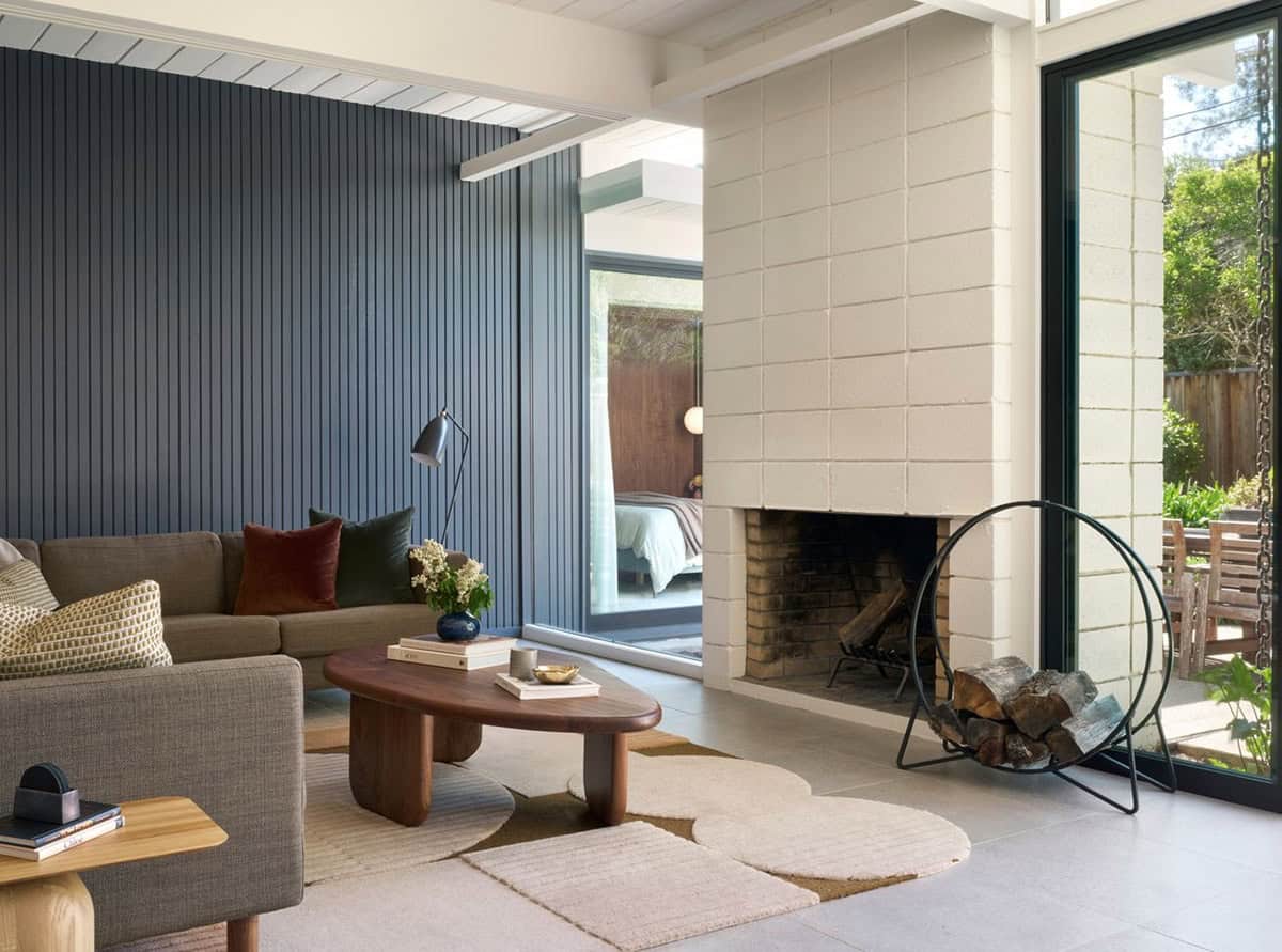 Living room with a gray sectional and walnut coffee table against a navy slatted wall, beside a white block fireplace and floor-to-ceiling glass