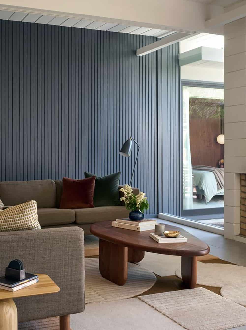 Gray sofa with velvet pillows, a walnut coffee table and a black floor lamp against a navy slatted wall, with a bedroom visible through the glass beyond
