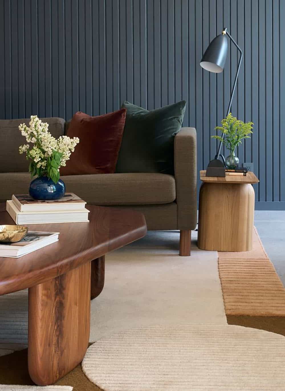 Walnut coffee table, olive sofa with rust and green velvet pillows, and a sculptural oak side table against a navy slatted wall