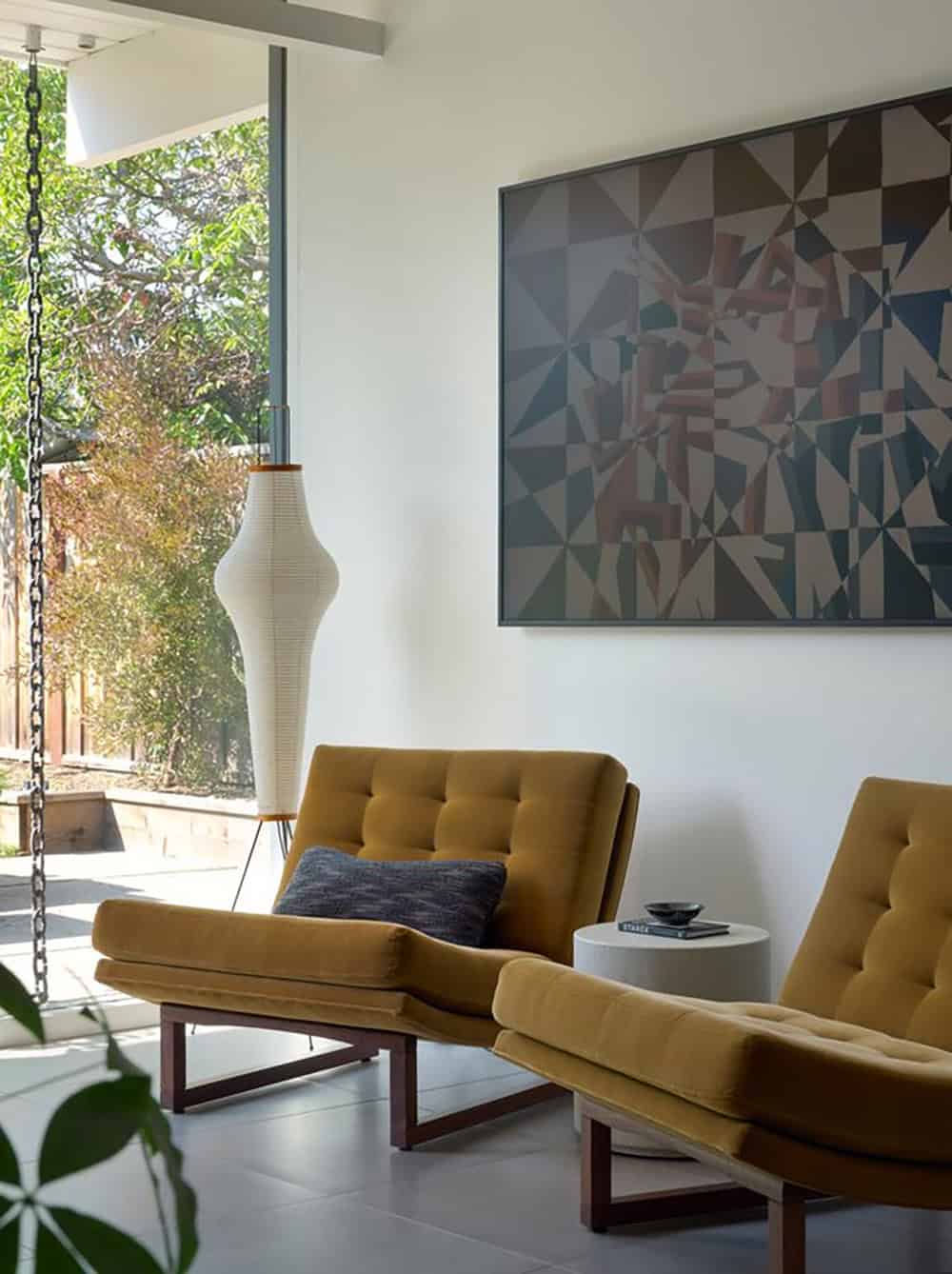 Two mustard velvet tufted lounge chairs beneath a large geometric painting, beside a paper floor lamp and a rain chain outside the glass