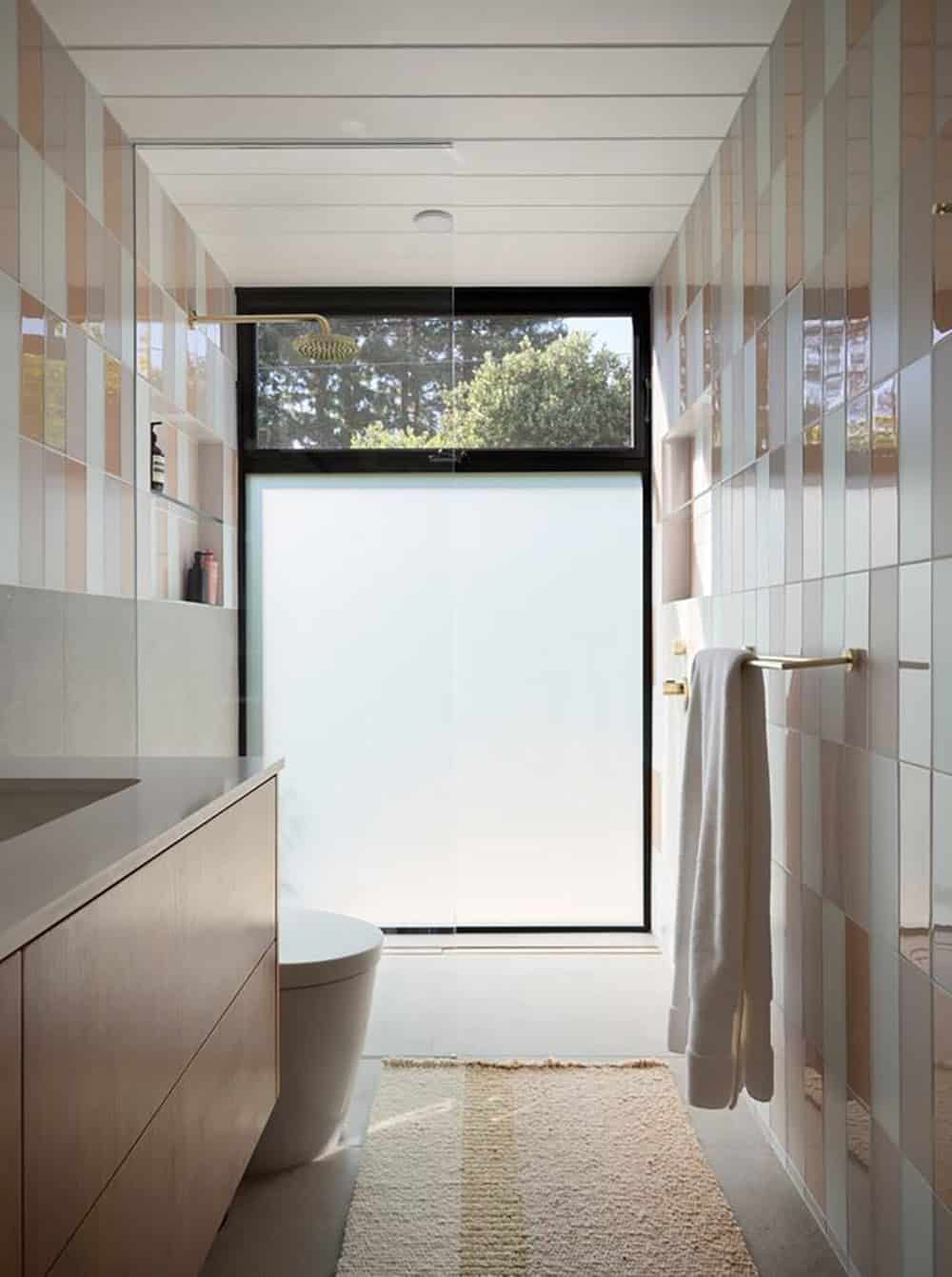 Curbless shower with stacked blush and white tile, a brass rain showerhead and a tall frosted window topped by a clear transom