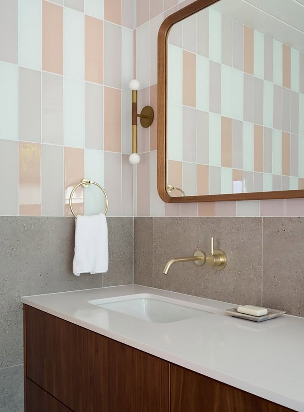 Brass sconce and towel ring above a speckled gray stone backsplash and walnut vanity, with blush and mint vertical tile above