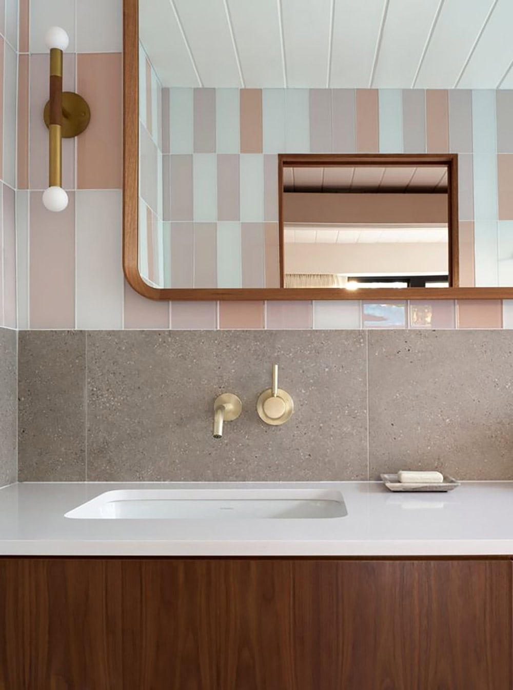 Wall-mounted brass faucet over an undermount sink against speckled gray stone, with a walnut-framed mirror and pastel tile above