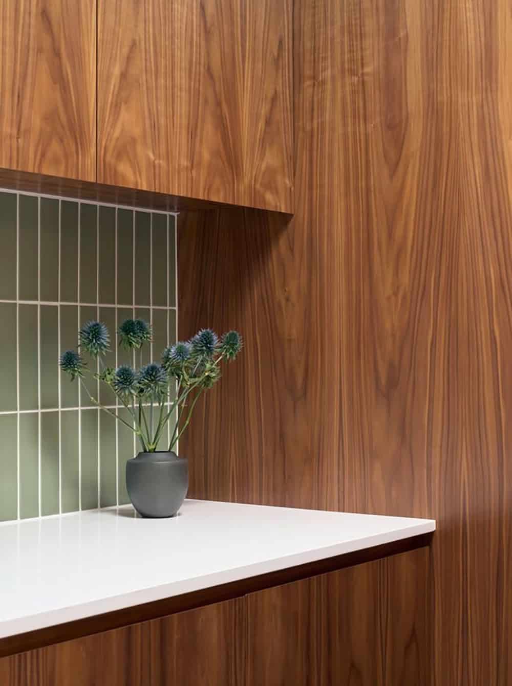 Walnut laundry room cabinetry with a white counter and sage green stacked tile backsplash, with a vase of blue thistles