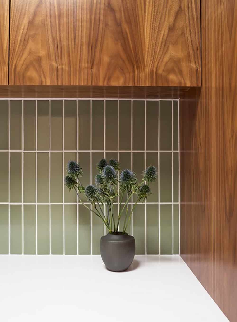 Sage green vertical stacked tile backsplash beneath walnut upper cabinets, with a dark vase of blue thistles on the counter
