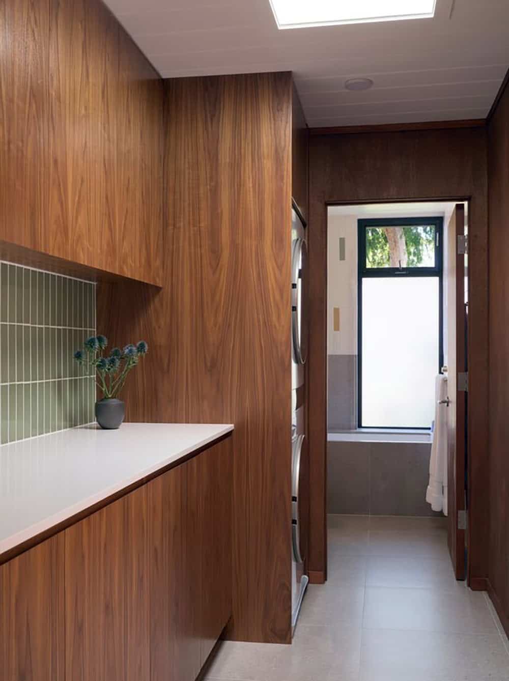 Walnut-paneled laundry room with a stacked washer and dryer, leading under a skylight to a bathroom with a frosted window