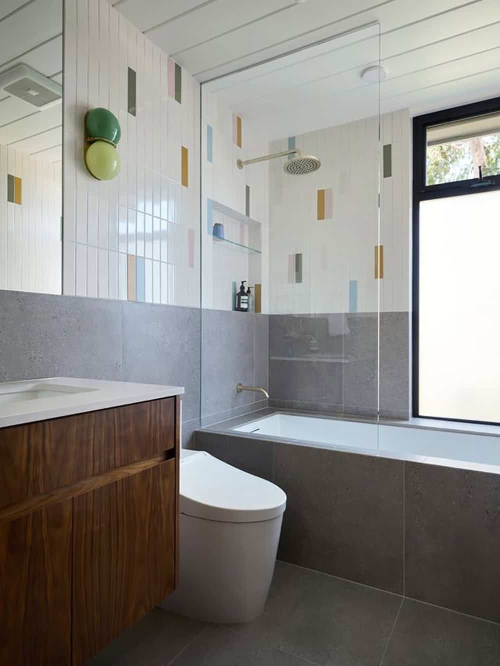 Bathroom with white vertical tile scattered with pink, blue, green and mustard accents, a walnut vanity, green globe sconce and tiled tub
