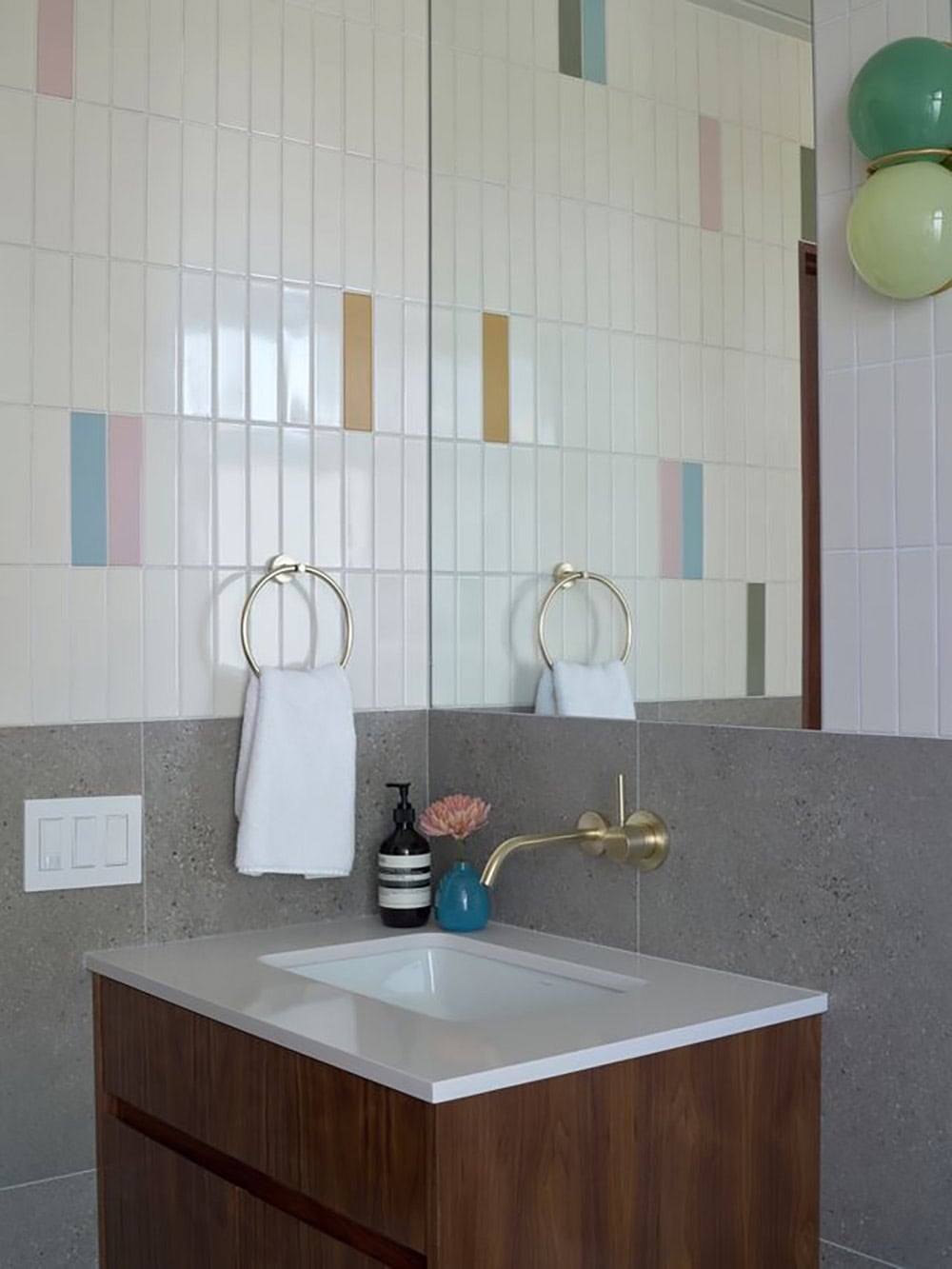 Walnut vanity with a brass wall-mounted faucet and towel ring against gray stone, below white tile dotted with pastel and mustard accents
