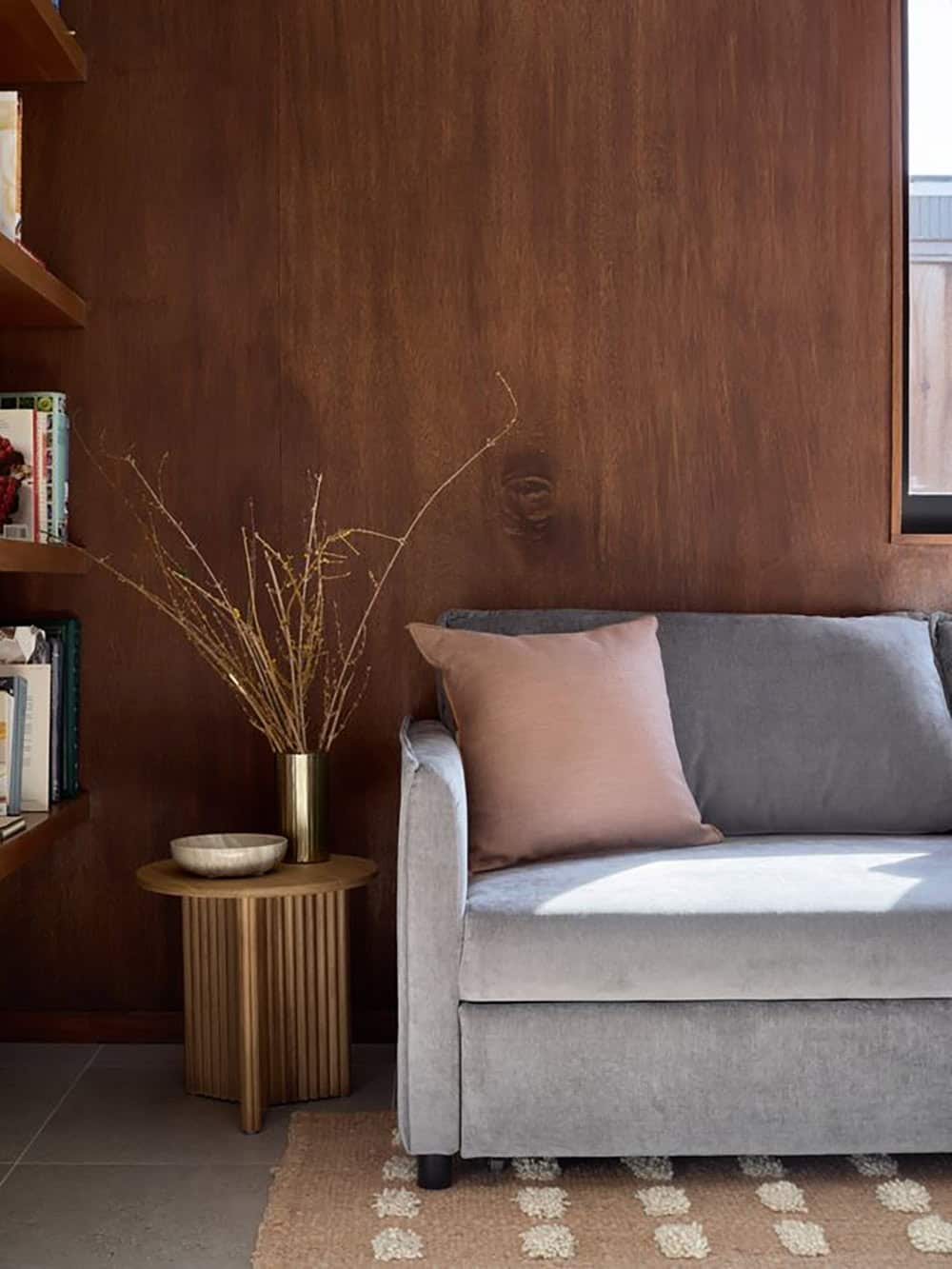 Gray velvet sofa with a blush pillow against a dark wood-paneled wall, beside a fluted wood side table, built-in bookshelves and a jute rug