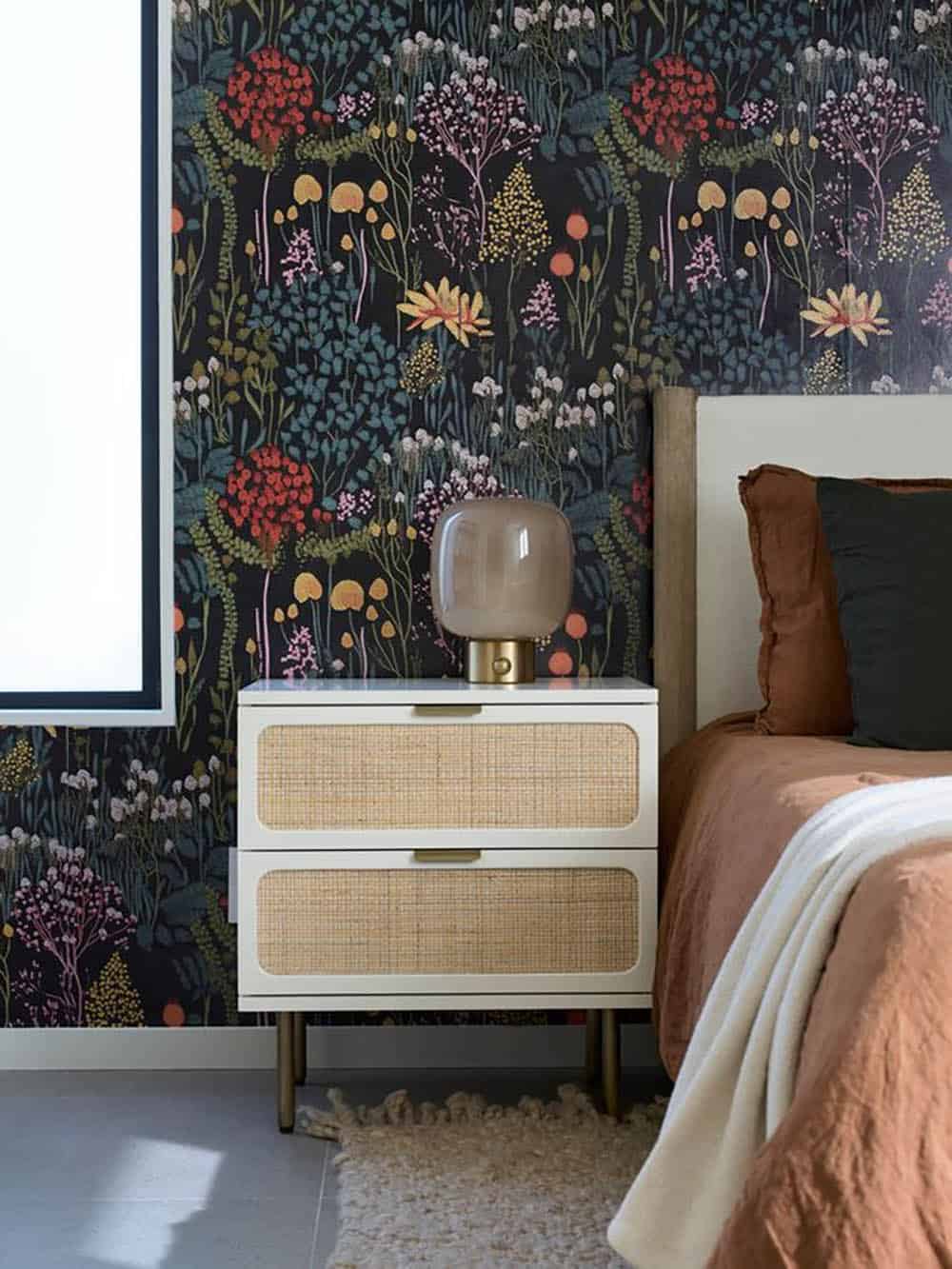 White and cane nightstand with a smoky glass lamp against dark wildflower wallpaper, beside a bed with rust linen bedding