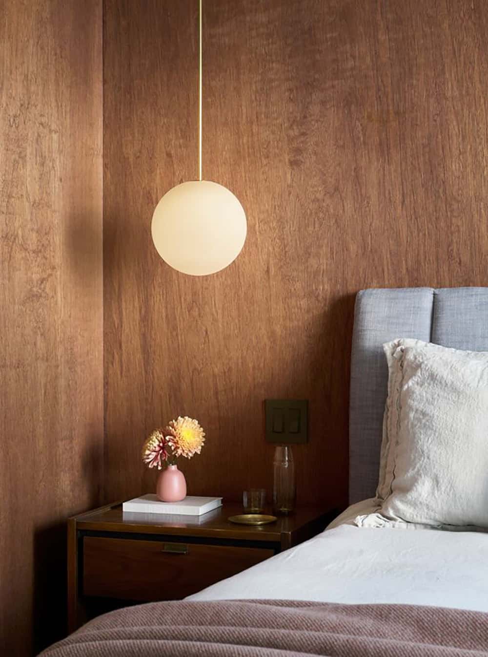 Warm-lit globe pendant hanging over a walnut nightstand against a wood-paneled wall, beside a blue-gray upholstered headboard