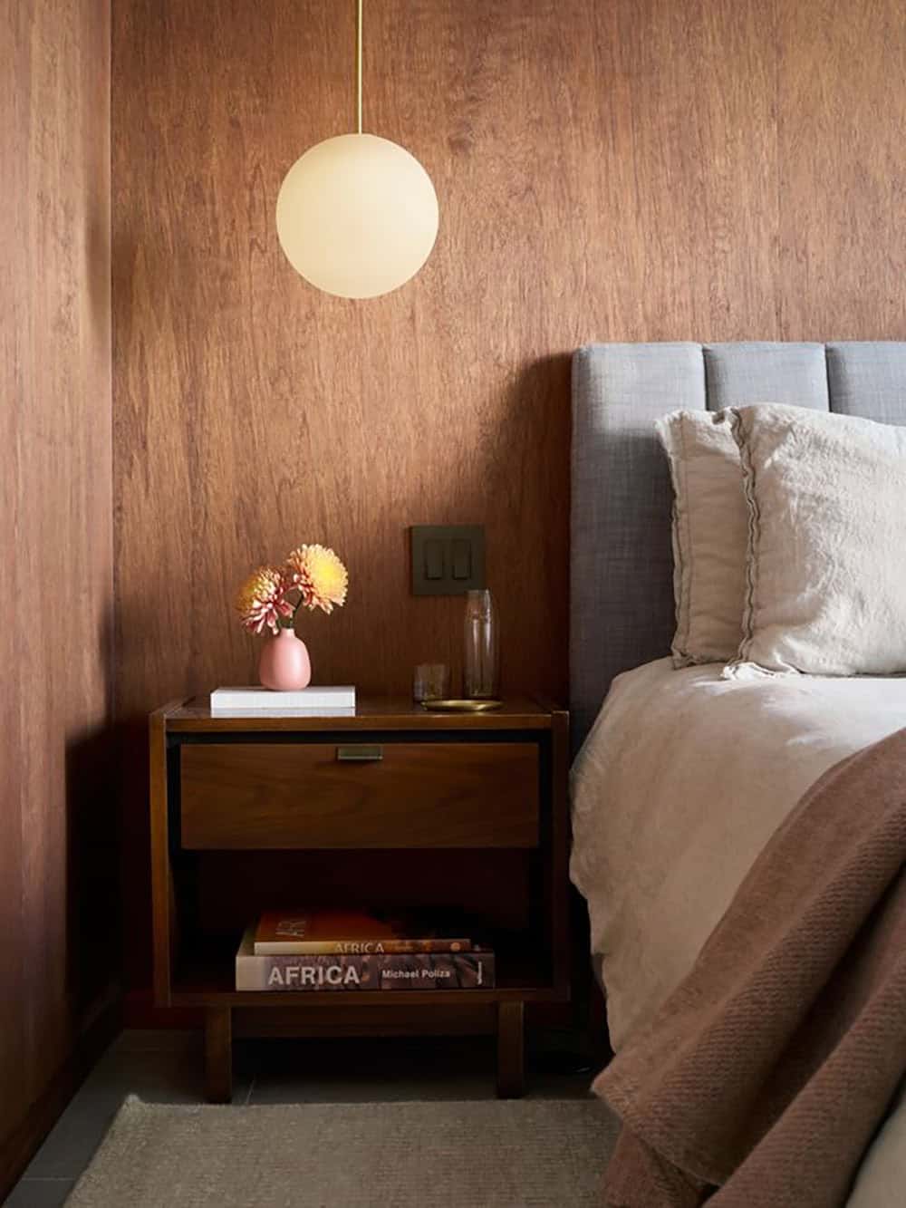 Vintage walnut nightstand with books on its lower shelf and a pink bud vase, under a warm-lit globe pendant against wood paneling