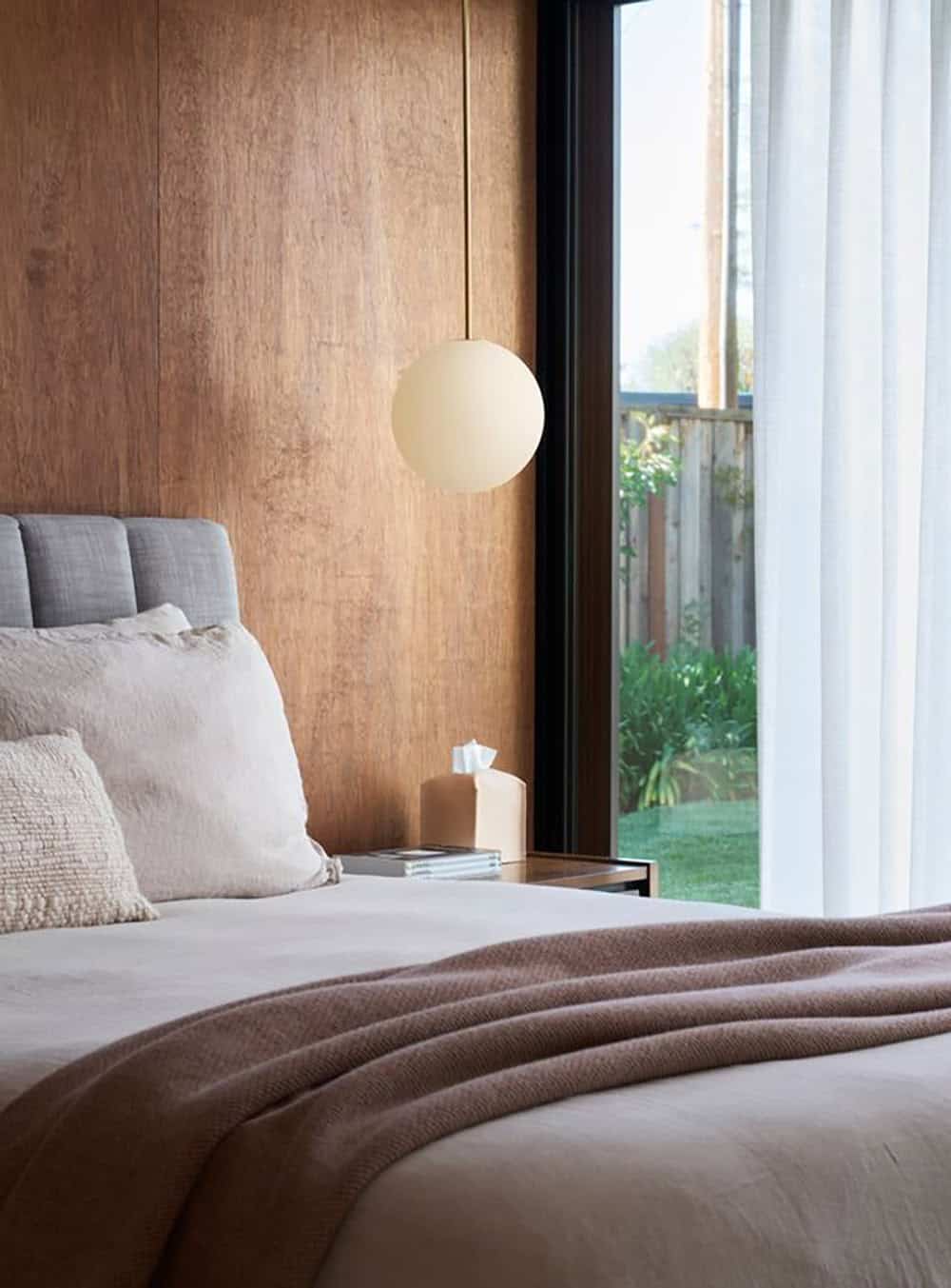 Bed with linen bedding and a mauve throw beside a warm-lit globe pendant and a corner window looking out to the garden