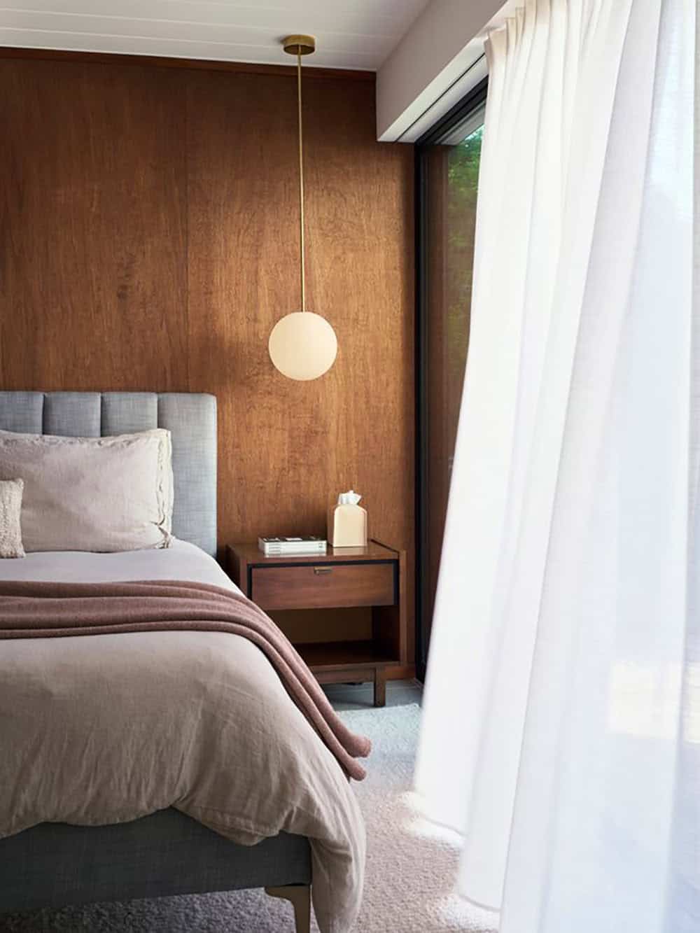 Channel-tufted bed and walnut nightstand under a warm-lit globe pendant, with sheer white curtains drawn across floor-to-ceiling glass