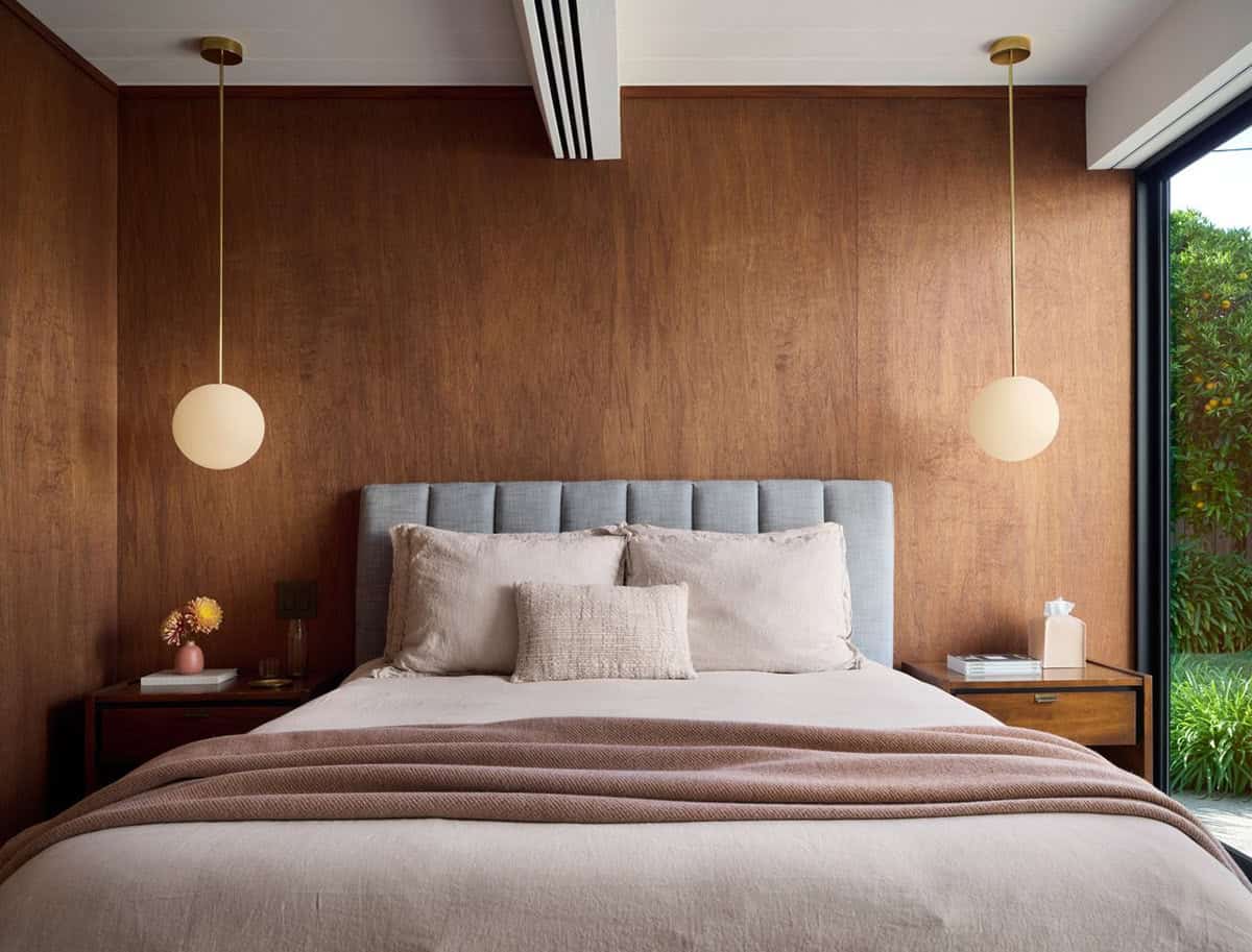 Bedroom with a blue-gray channel-tufted bed centered on a wood-paneled wall between two brass globe pendants casting warm light, with glass to the garden