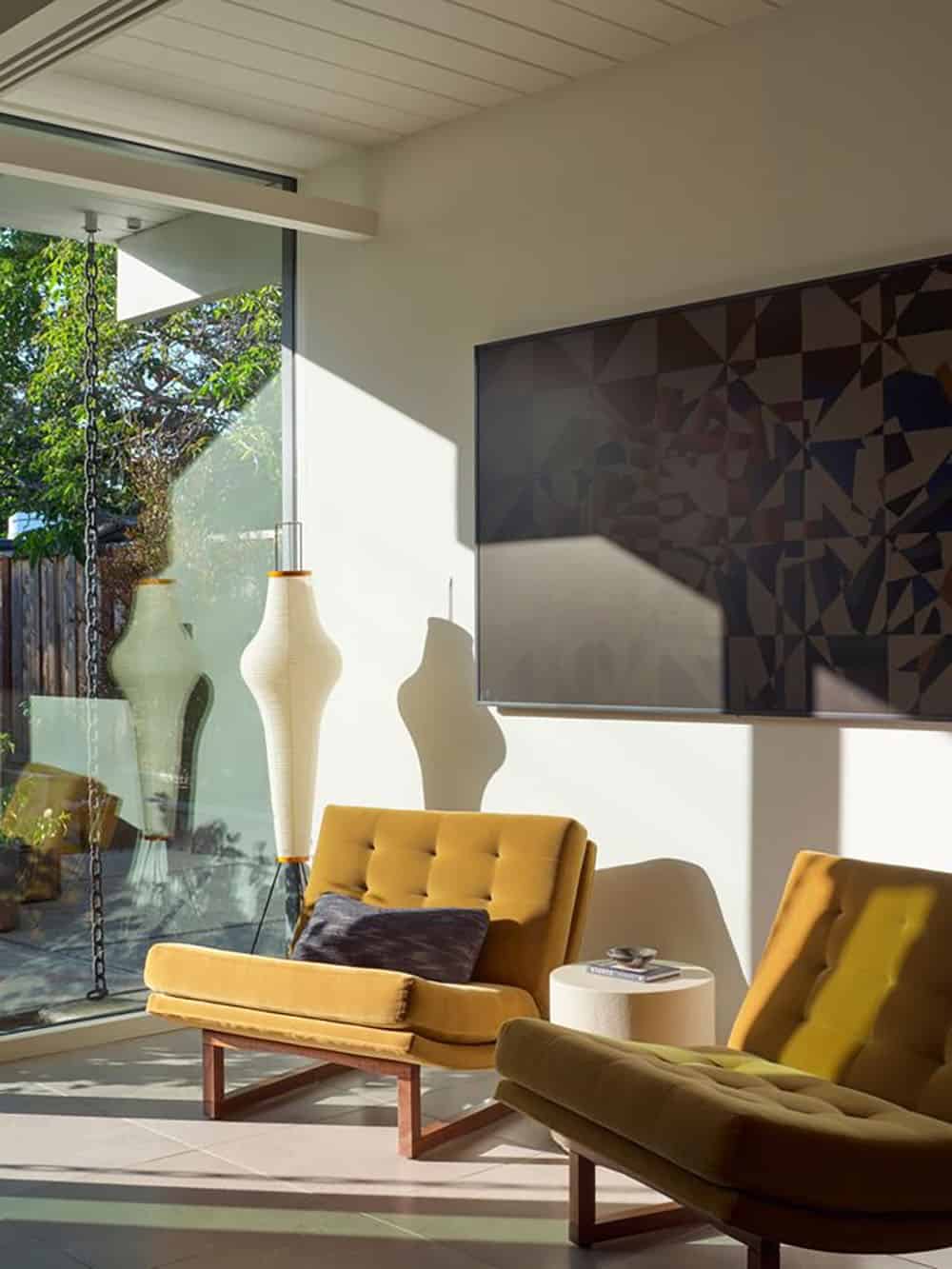 Sunlit sitting area with mustard velvet lounge chairs, a paper floor lamp and a geometric painting, with a rain chain outside the glass