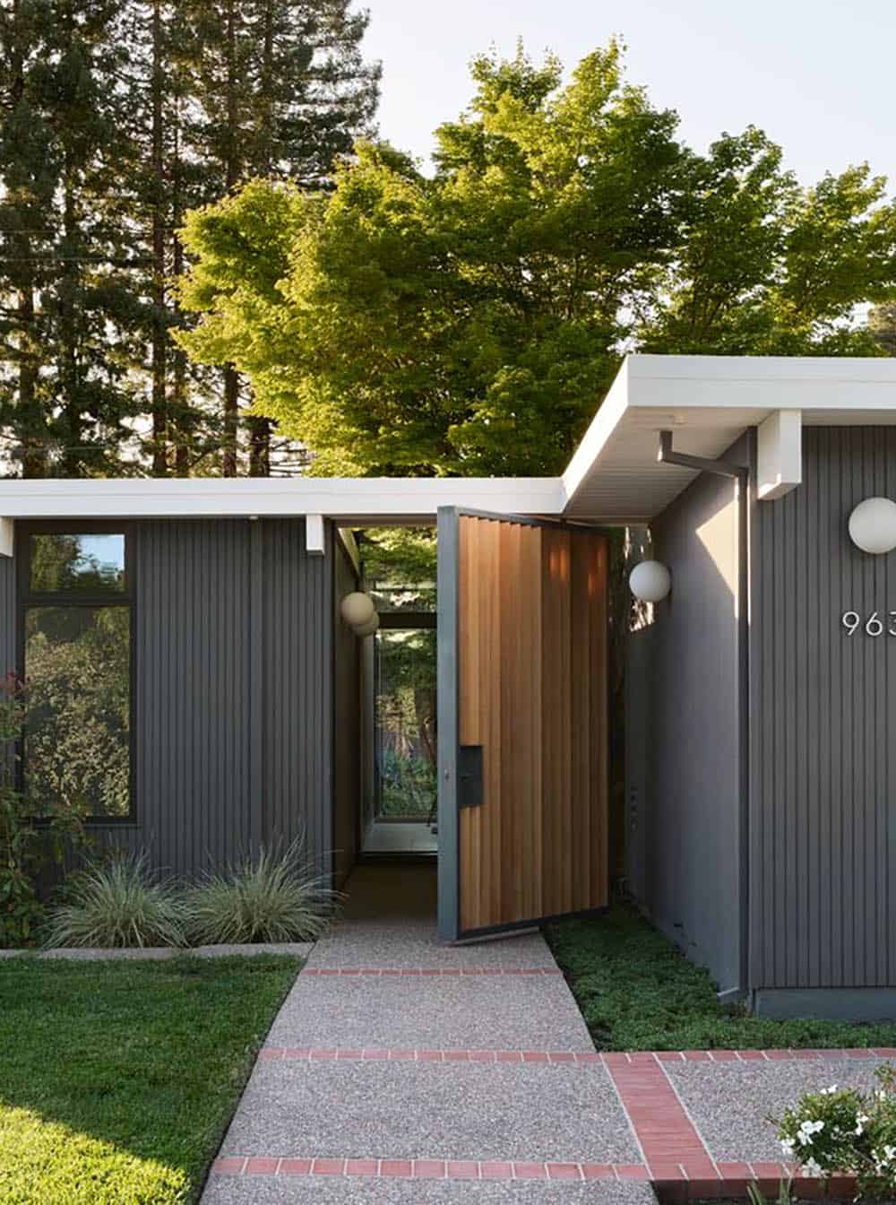 Eichler front entry with dark gray vertical siding and a slanted cedar slat pivot gate opening onto an exposed aggregate walkway with brick bands