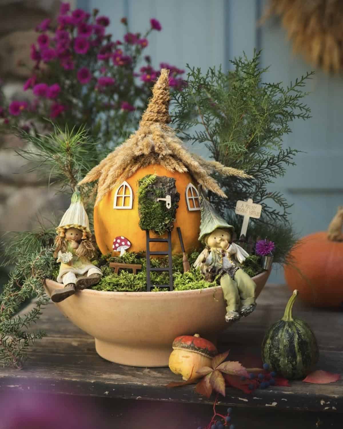 no-carve pumpkin fairy house with thatched grass roof, moss door, and fairy figurines in a terra-cotta bowl