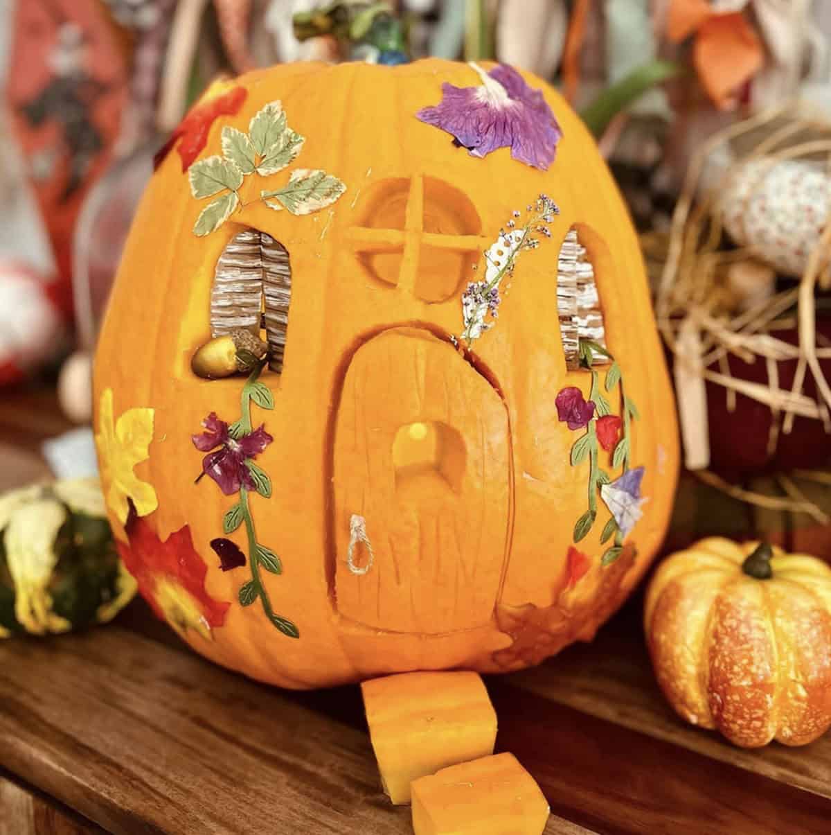 orange jack-o’-lantern with carved arched door, paper window curtains, pressed flowers, and climbing vines