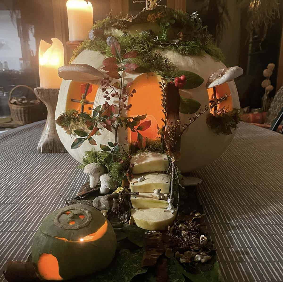 white fairy house pumpkin with glowing windows, moss roof, shelf mushrooms, berry branches, and porcelain cats on pumpkin steps