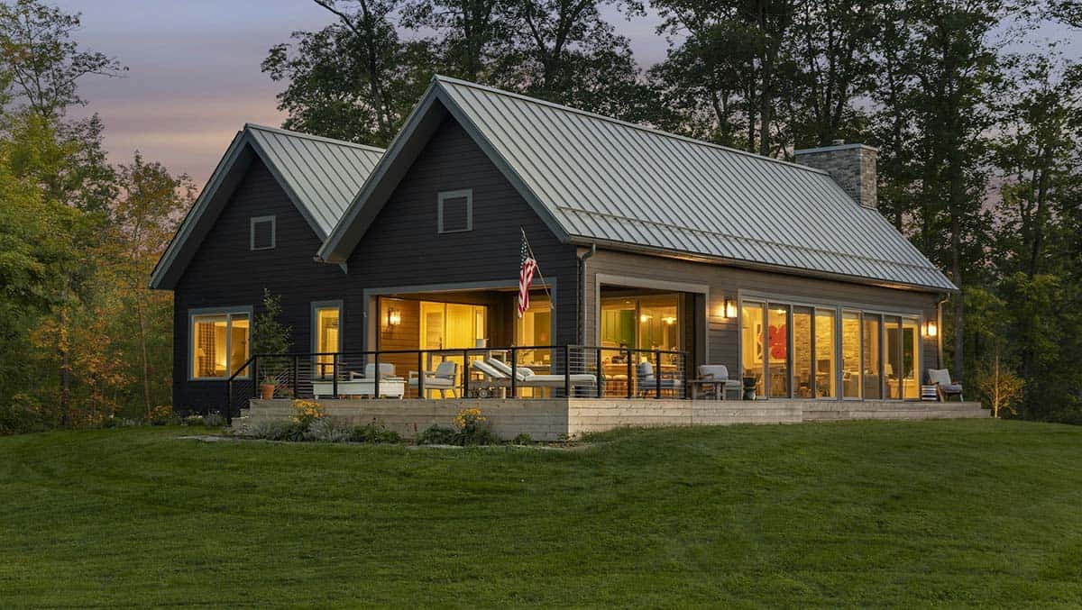 Dark gray gabled lake cabin with a metal roof glowing at dusk, with a covered porch, cable-rail deck and a wall of glass doors