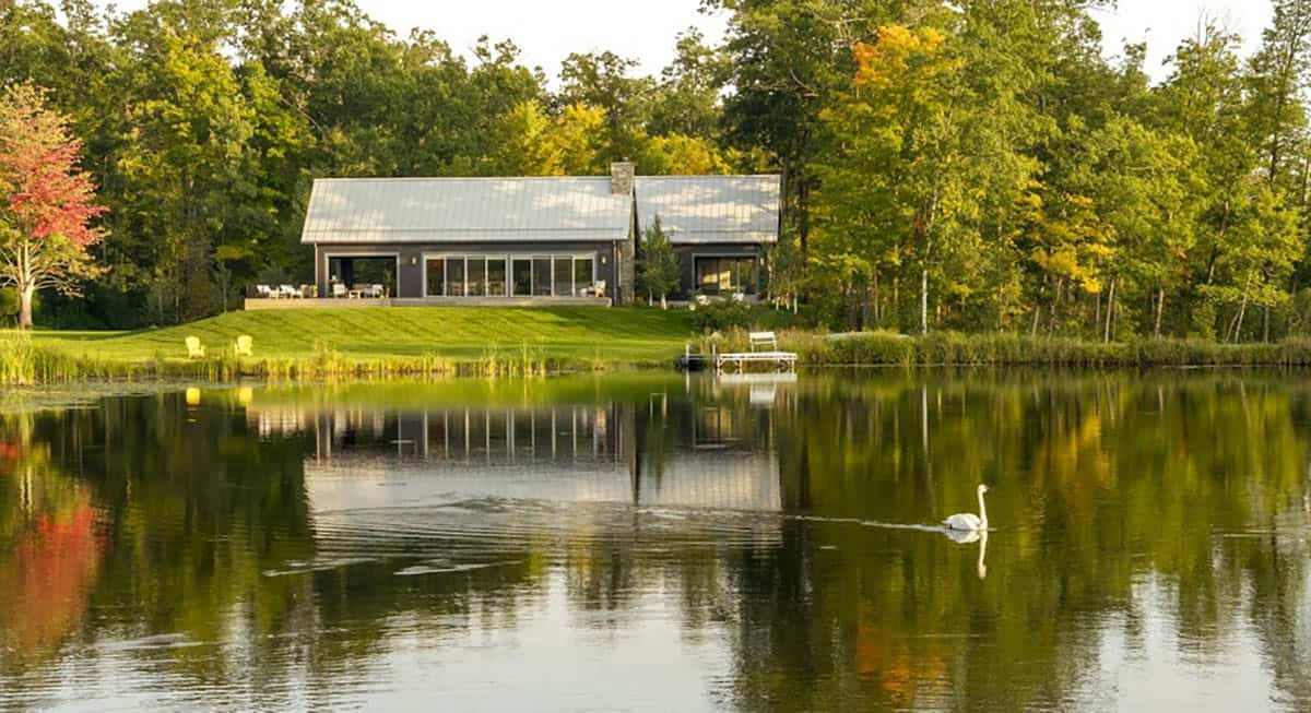 Dark gray cabin with two metal-roofed gables reflected in a calm lake, with a swan, a small dock and early fall trees