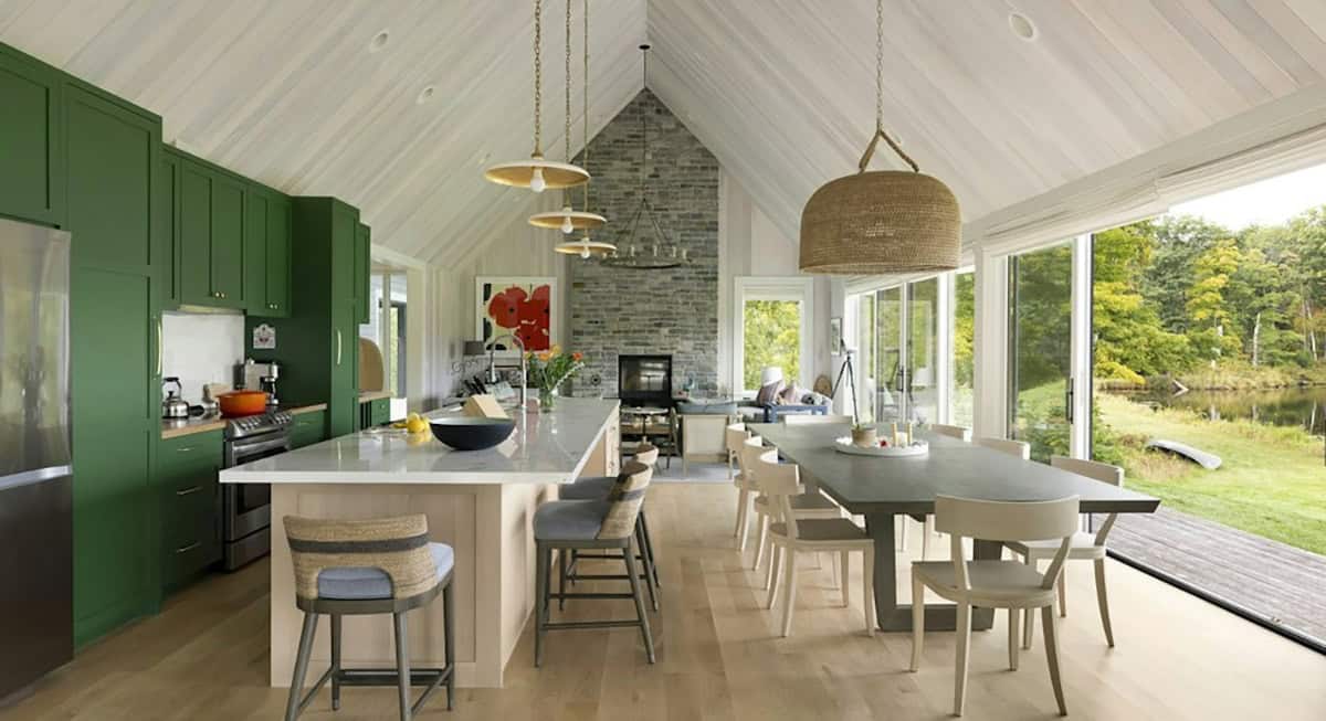 Open kitchen with green cabinets, a white oak island with woven-back stools and a long dining table by glass doors to the lake, under a vaulted whitewashed ceiling